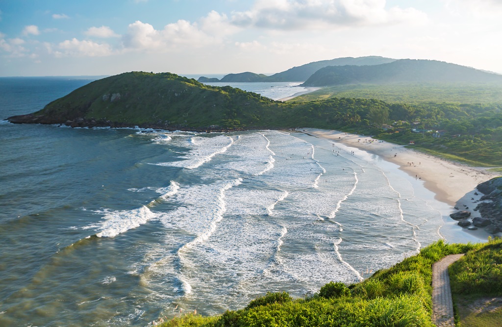 Vista aérea da Praia de Fora e Praia de Dentro na Ilha do Mel, separadas por uma faixa de areia e o morro verde, com mar agitado.
